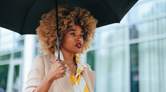 Close up shot of a beautiful african american woman holding a black umbrella and walking down the street. She is smiling while looking away.