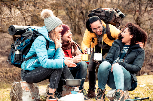 Happy multiracial friends sitting drinking cup of coffee after a day of hiking in the mountains - Traveler people camping in the forest and relaxing together - Concept of adventure trekking vacation