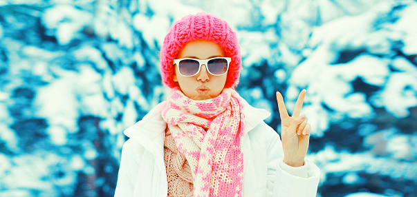 Fashion winter portrait of stylish young woman wearing sunglasses, colorful knitted hat and scarf in the park on snowy background