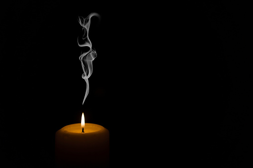 A flame of a burning white wax candle with smoke emanating and rising on a black background