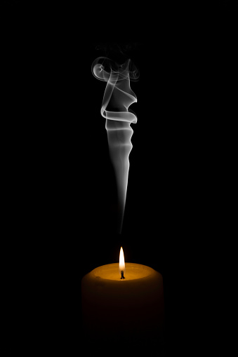 A flame of a burning white wax candle with smoke emanating and rising on a black background