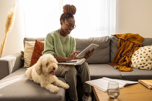 Focused cute stylish African American female student, studying remotely from home, digital tablet, taking notes on notepad during online lesson, e-learning concept. She has a beautiful pet dog by her side