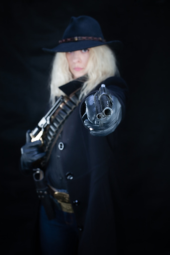 wild west blonde girl wearing black hat shooting from revolver on black background