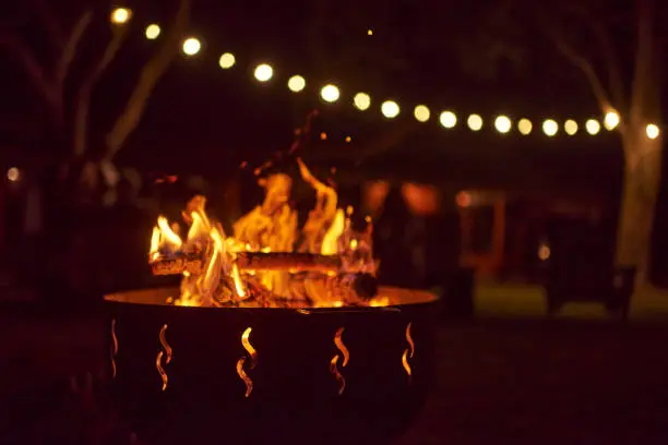 Bonfire burning in a garden on a warm summer night, unrecognizable people in the background. Concepts: coziness, travel, enjoyment of the outdoors.