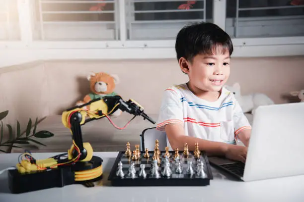 Photo of Asian little boy programming code to robot machine arm on laptop for play chess