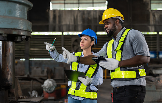 African American male and female engineers in safety vest and helmet checking and repairing old machine at heavy metal industrial factory. Preventive Maintenance concept