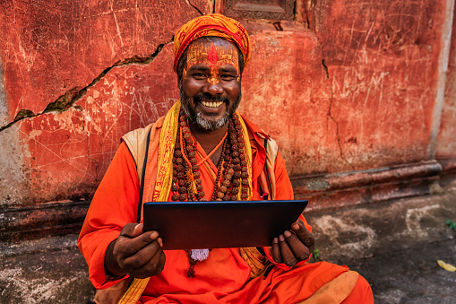 Sadhu - indian holyman using the digital tablet. In Hinduism, sadhu, or shadhu is a common term for a mystic, an ascetic, practitioner of yoga (yogi) and/or wandering monks. The sadhu is solely dedicated to achieving the fourth and final Hindu goal of life, moksha (liberation), through meditation and contemplation of Brahman. Sadhus often wear ochre-colored clothing, symbolizing renunciation.