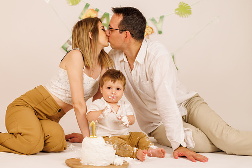 Happy family celebrating baby's first birthday with birthday cake indoor, parents kissing while boy eating birthday cake with hands