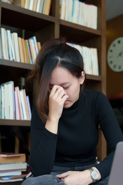 istock A portrait of a young Asian woman using a computer, wearing headphones and using a notebook to study online shows boredom and pain from video conferencing on a wooden desk in library 1436016819