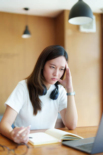 istock A portrait of a young Asian woman using a computer, wearing headphones and using a notebook to study online shows boredom and pain from video conferencing on a wooden desk in library 1436017989