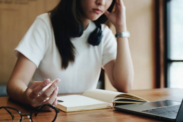 istock A portrait of a young Asian woman using a computer, wearing headphones and using a notebook to study online shows boredom and pain from video conferencing on a wooden desk in library 1436018012