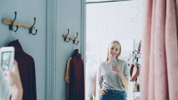 istock Pretty blond woman is making mirror selfie with smart phone while standing in fitting room in clothes boutique. She is posing, moving and smiling carelessly. 1436018052