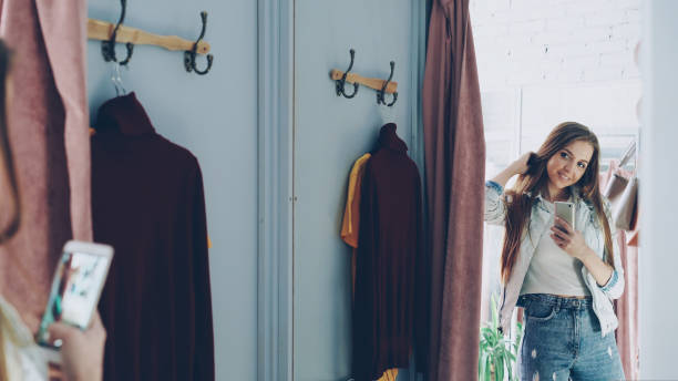 istock Pretty girl is making mirror selfie with smartphone while standing in fitting room in clothes store. She is posing, turning and touching her hair with lovely smile. 1436018125