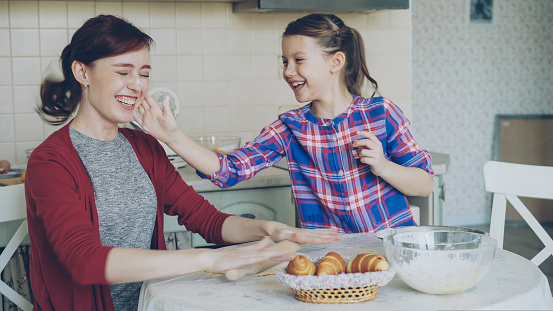 Young mother and her cute daughter have fun smearing nose each other with flour while cooking together in the kitchen on holidays. Family, food, home and people concept