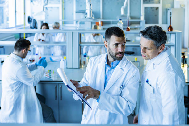 Mid adult male chemists talking while working on scientific research in laboratory. Male experts cooperating while going through medical data in laboratory. There are people in the background. biochemist stock pictures, royalty-free photos & images