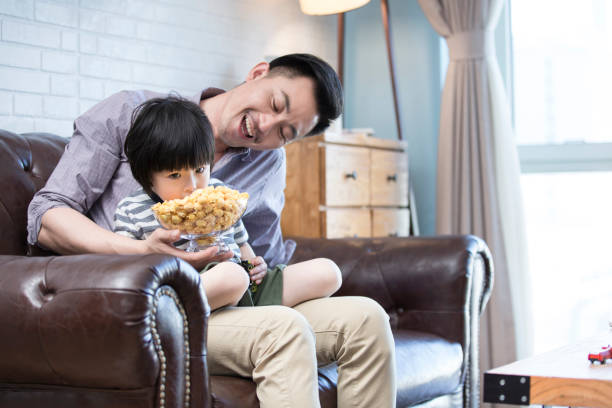 Middle-aged father sitting on the family living room sofa holding his son and feeding him popcorn - stock photo Suitable for family education, parent-child, preschool education, children's interest development, intellectual development, hobby development, soccer parent-child, sports exercise, Father's Day, Mother's Day, Children's Day, holidays, family entertainment, children's nutrition, soccer World Cup, soccer tournament, print advertising. boys bowl haircut stock pictures, royalty-free photos & images