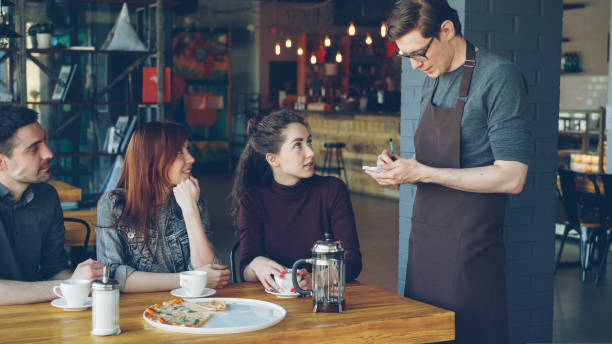 friendly male waiter is taking order from cheerful group of friends sitting at table in cafe and talking. eating out, food and drink industry and friendship concept. - restaurant food food and drink industry food service occupation imagens e fotografias de stock