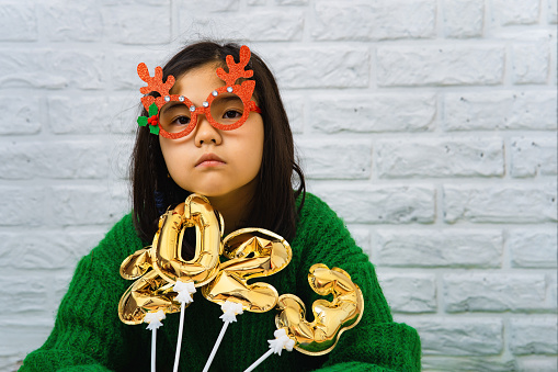 Sad asian child girl in green sweater in masquerade christmas glasses with balls with number 2023. Portrait of vietnamese girl in masquerade glasses with deer horns, concept of christmas masquerade, party