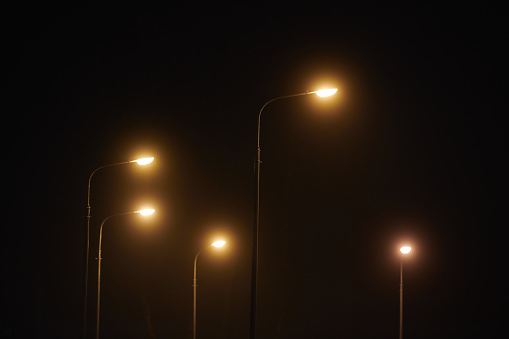 Night lampposts shines with faint mysterious yellow light through evening fog. Streetlights shine at quiet city night, magic atmospheric light in mystical darkness