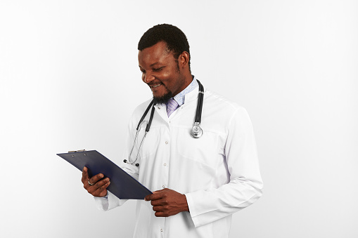 Smiling black bearded doctor man in white robe with stethoscope looks to medical chart on clipboard, isolated on white background. Happy adult black african american physician therapist portrait