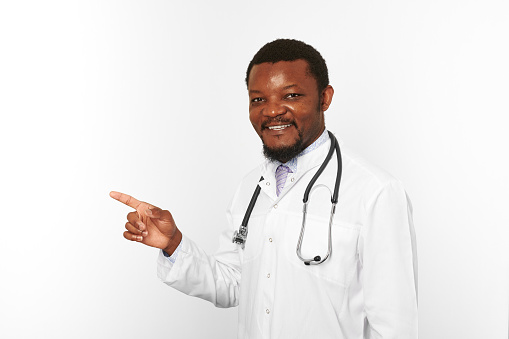 Smiling black bearded doctor man in white coat with stethoscope points finger to left, isolated on white background. Adult black african american physician therapist portrait, candid male emotion