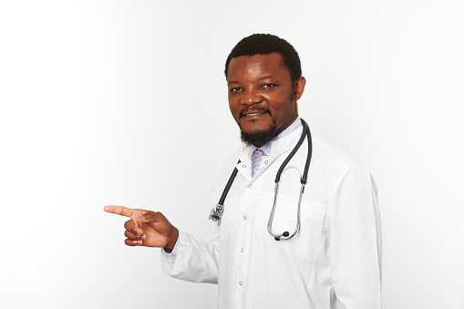Smiling black bearded doctor man in white coat with stethoscope points finger to left, isolated on white background. Adult black african american physician therapist portrait, candid male emotion