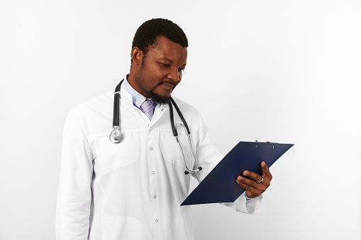 Black bearded doctor man in white robe with stethoscope looks to medical records on clipboard, isolated on white background. Serious adult black african american physician therapist portrait