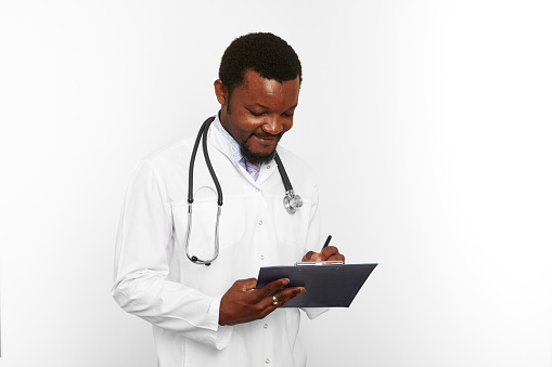 Black bearded doctor man in white coat with stethoscope filling medical records on clipboard, isolated on white background. Thoughtful adult black african american physician therapist portrait