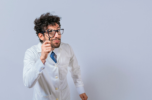 Mad scientist holding a magnifying glass isolated, Man in white coat with magnifying glass looking at camera, Surprised male scientist in lab coat looking at camera with magnifying glass
