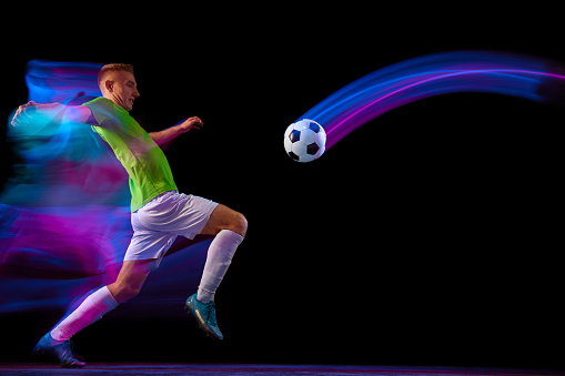 Sport in action. One man professional soccer player training with football ball isolated on dark background in neon mixed light. Sport, speed, power and energy. Energetic athlete practicing
