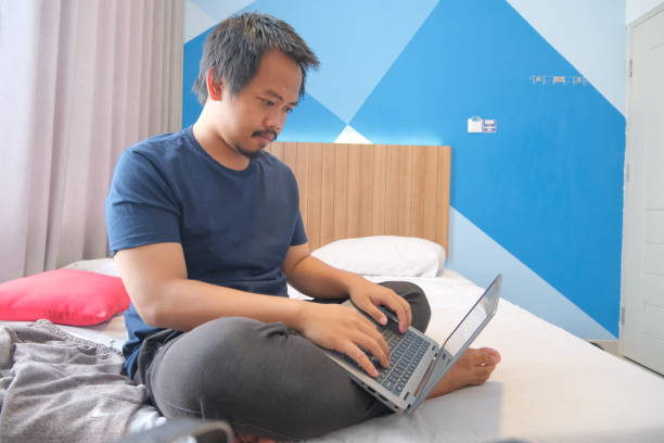 mężczyzna z azji południowo-wschodniej pracujący zdalnie z łóżka hotelowego - southeast asian ethnicity men laptop image type zdjęcia i obrazy z banku zdjęć