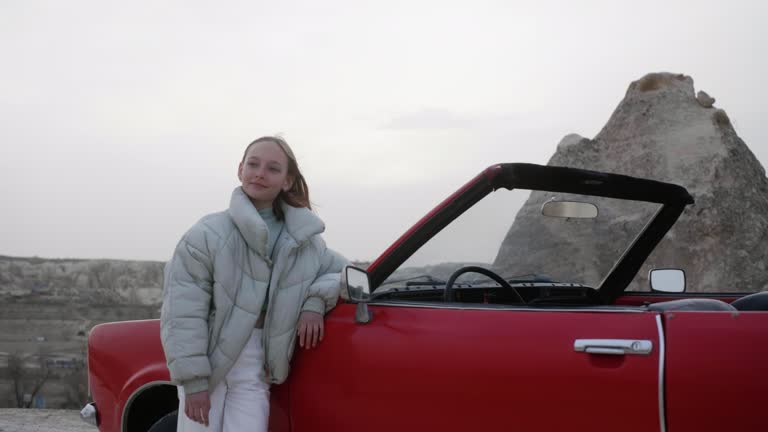 Attractive teen girl in green jacket stand near red loan cabrio car and enjoy landscape desert stone valley of Cappadocia in Turkey. Zoom out.