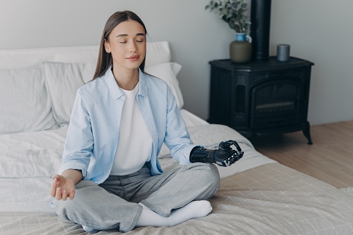 Calm young disabled girl with bionic hand prosthesis practicing yoga sitting on bed at home. Female with artificial arm limb meditates folded fingers in mudra gesture. Disability and healthy lifestyle