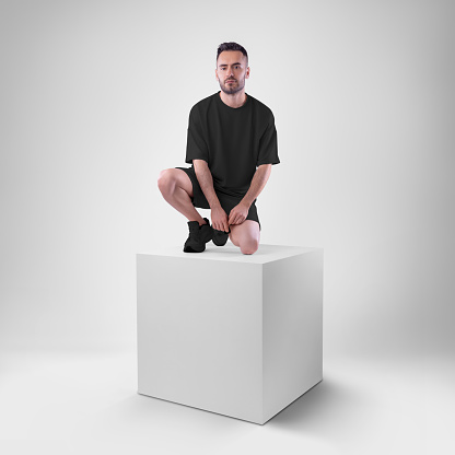 isolated mockup of trendy black men's oversized t-shirt with shorts on man sitting on cube. Clothing template for presentations of design, print, pattern.