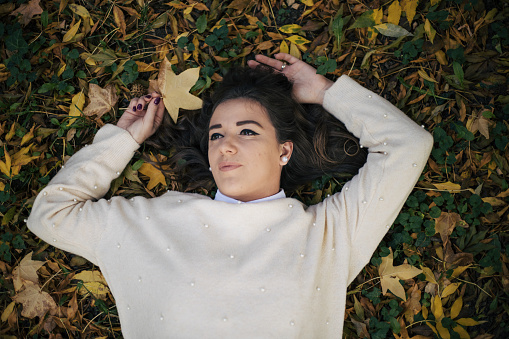 Young woman with curly brown hair posing in nature.In white sweather and autumn clothing style. Beautiful colored leafes and idyllic atmosphere. Holding leafes while Lzing donw on the grass.