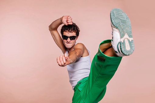 Portrait of handsome guy, throwing high kick in the air, practicing self defense kicking confident facial expression wear sun specs, white t-shirt and green pants isolated over beige color background.