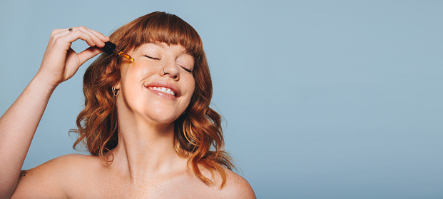 Happy woman with ginger hair applying face serum using a dropper in a studio. Cheerful young woman treating her skin with cosmetic oil. Smiling woman applying a nourishing beauty product on her face.
