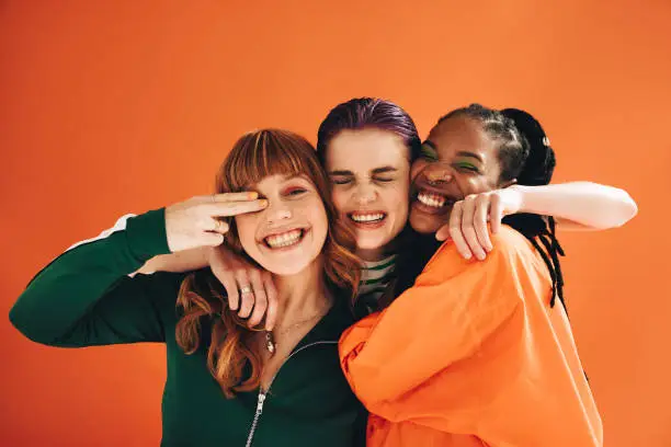 Three multicultural female friends smiling and embracing each other in a studio. Group of vibrant young women enjoying themselves while standing against an orange background.