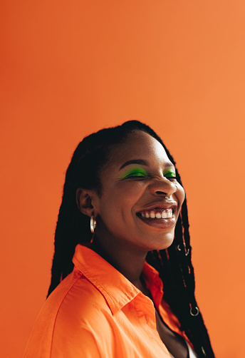 Cheerful african woman with makeup and face piercings smiling with her eyes closed. Happy young woman with dreadlocks standing against a studio background. Black female hipster feeling confident in her style.
