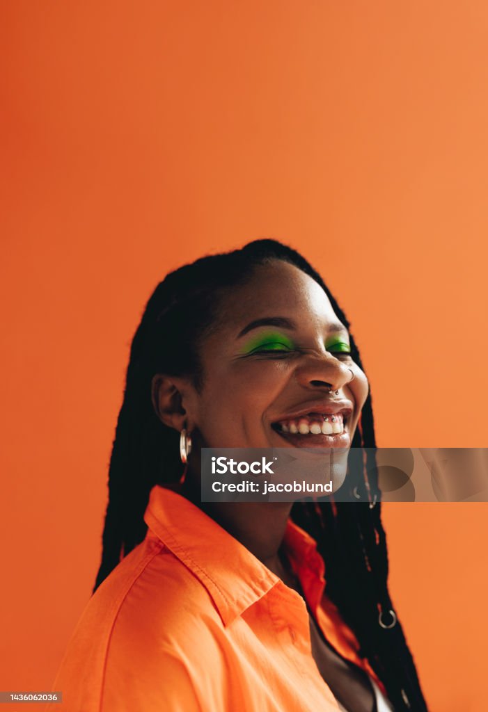 Cheerful african woman with makeup and face piercings smiling in a studio Cheerful african woman with makeup and face piercings smiling with her eyes closed. Happy young woman with dreadlocks standing against a studio background. Black female hipster feeling confident in her style. One Woman Only Stock Photo