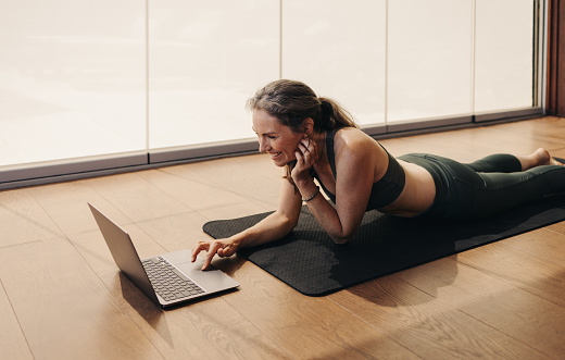 Elderly woman smiling while joining a virtual fitness class on a laptop. Happy senior woman following an online yoga tutorial at home. Cheerful mature woman lying on an exercise mat.