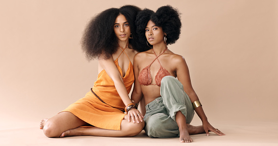Portrait of black women sisters in studio with natural beauty, fashion and style. Young female model with sister sitting on floor with stylish outfit for summer. Beautiful females with afro hairstyle