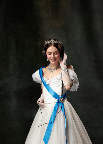 Favorite music. Elegant woman, royal person, queen or princess in headphones and medieval outfit listening track on vintage background. Concept of comparison of eras, modern sweets, fashion, beauty, ad
