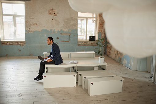 Portrait of young businessman sitting in empty room and working on laptop on new business project. Opening new company. Success. Concept of business, challenges, work, occupation, professional growth