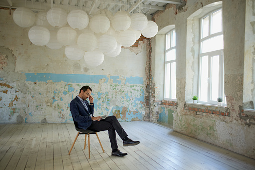 Portrait of young thoughtful man, businessman sitting on chair with laptop in empty room. Choosing building for company. Concept of business, challenges, work, occupation, brainstorming