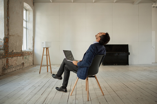 Portrait of young businessman sitting in empty room with laptop and looking at ceiling. Business failure, professional difficulties. Concept of business, challenges, work, occupation