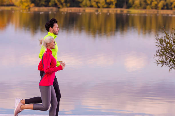 running couple while training outdoors on road near river bank for durable marathon pretty young female and extremely fit caucasian handsome man in their prime. - pair couple mid adult happiness imagens e fotografias de stock