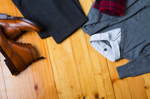 View Flat Lay of Tan Brogues Boots With Jumper, White Shirt,  Sweater, Pair of Herringbone Trousers on Wooden Surface Background.Horizontal image
