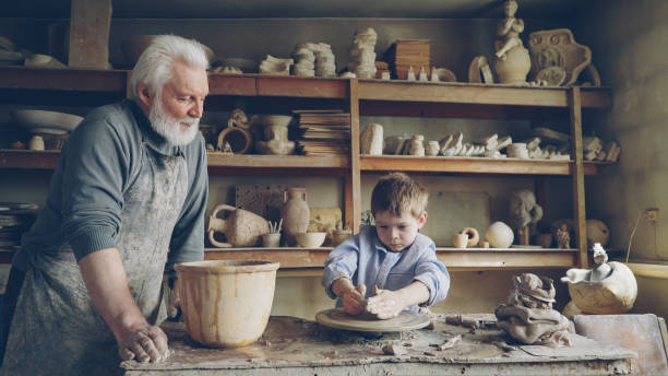 istock Small boy is molding clayware in his grandfathers's studio while his caring senior grandpa is standing near him and watching him with love. Family traditions concept. 1436082987