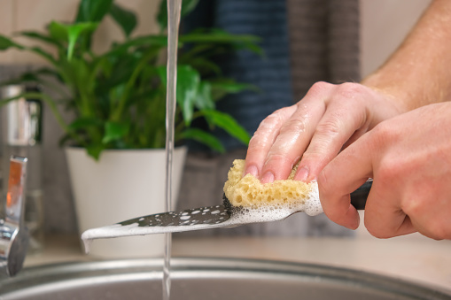A man washes a sharp steel dirty kitchen knife with a sponge with detergent in the kitchen sink under running water. Gentle hand washing of the kitchen knife. Cleaning of dirty sharp metal products.